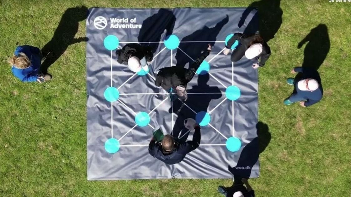 På billedet ses en gruppe deltagere i gang med teambuilding-aktiviteten "Optimizer". De står samlet omkring en stor måtte med et netværksmønster af blå felter og forbindelseslinjer. Opgaven kræver samarbejde, strategi og kommunikation for at finde den mest optimale vej gennem mønsteret. Aktiviteten foregår udendørs i solskin og stimulerer både logisk tænkning og teamwork.