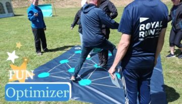 Billedet viser en gruppe deltagere samlet omkring en stor måtte med forbundne punkter i et netværksmønster – et spil kaldet "Optimizer". Aktiviteten foregår udendørs og kræver samarbejde, strategi og kommunikation for at finde den optimale løsning. Det er en sjov og udfordrende teambuilding-øvelse, hvor deltagere skal tænke logisk og arbejde effektivt sammen.