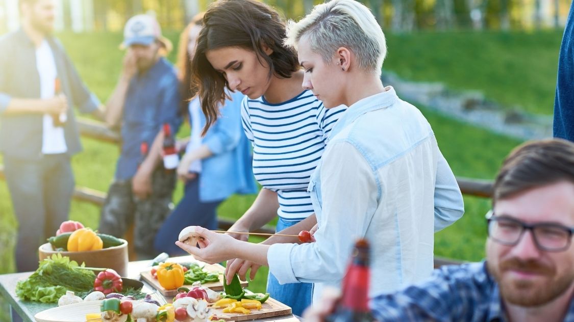 Billedet viser en udendørs teambuildingaktivitet, hvor deltagerne forbereder mad sammen i det fri. To personer arbejder koncentreret med at lave grillspyd af friske grøntsager, mens andre nyder stemningen i baggrunden. Aktiviteten lægger op til samarbejde, kreativitet og hygge – perfekt til sommerarrangementer eller firmafester under åben himmel.