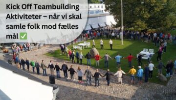 står i en stor cirkel og holder hinanden i hånden, hvilket skaber en stærk følelse af fællesskab og samarbejde. Arrangementet foregår på en grøn plæne foran en moderne bygning med store glaspartier og hvide facader. Stemningen virker positiv og engageret, og det ser ud til at være en del af et Kick Off-event. I forgrunden er der brosten og nogle store sten, og vejret er klart med solskin, hvilket bidrager til en behagelig atmosfære.